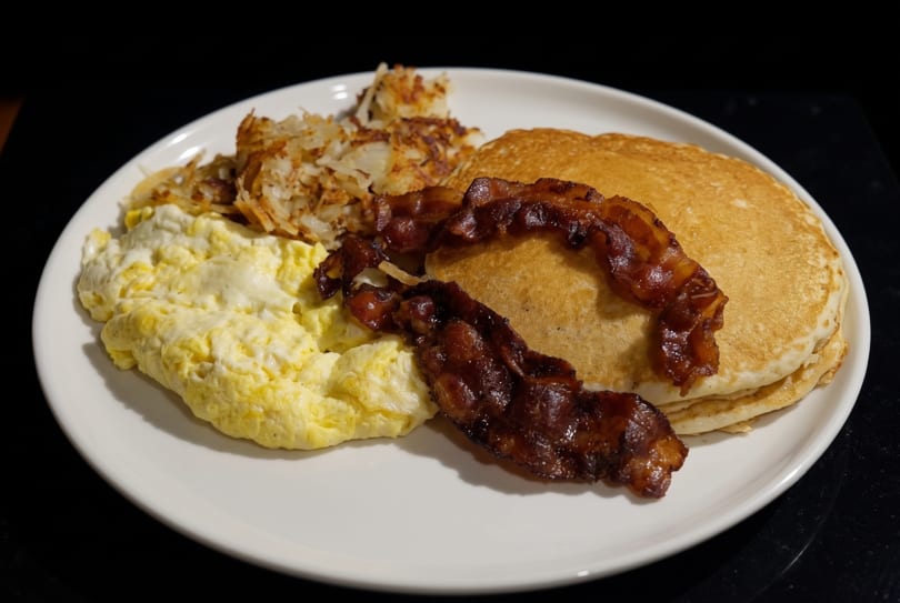 A breakfast plate with scrambled eggs, crispy hash browns, bacon strips, and a stack of pancakes.