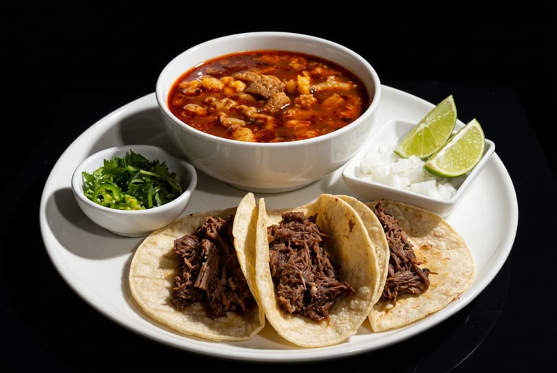 Three beef tacos, a bowl of menudo soup, and sides of cilantro, jalapeños, onions, and lime.